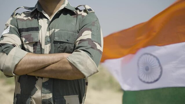 Indian soldier standing with arms crossed in front of waving indian flag - concept of confident, fearless and pride serviceman
