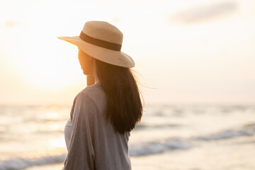 Young asian woman with straw hat standing alone on empty sand beach at sunset seashore. Chilling in holiday weekend summertime. Traveler Female walking around the beach with the sunlight