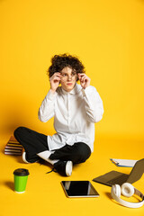 Portrait of young man, student in glasses sitting on floor around books, laptop and tablet isolated over yellow studio background