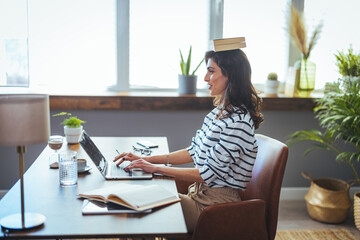 Shot of young attractive woman at the desk with books on her head while working with computer at...