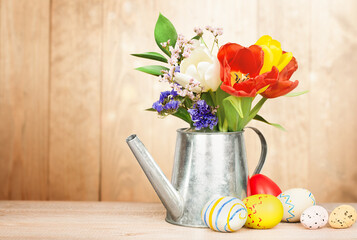 spring flowers in a decorative watering can and easter eggs on a wooden background