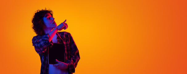 Portrait of young curly man in checkered shirt with tablet pointing right side with interest isolated over orange background in blue neon