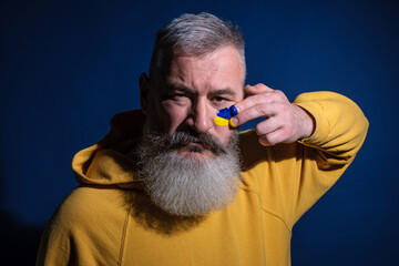 Mature bearded man dressed yellow hoodie draws Ukrainian flag on his face, protest against war