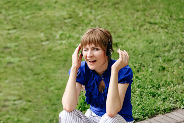 Fototapeta premium Grimacing Girl Listening Music through Headphones. Happy and Attractive Young Woman with Earphones Listen Compact Audio Player in Park. Female with Court Brunette Hair Wearing Blue Blouse