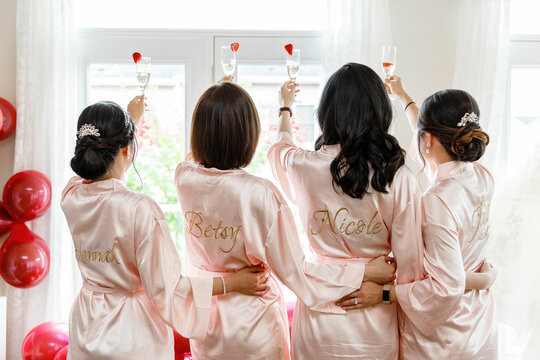 Bridesmaids and bride in pink robes standing with their backs to the camera holding champagne glasses  and hugging Back view