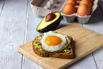 Whole grain toast with mashed avocado and fried egg on wooden white table. Tasty healthy breakfast.