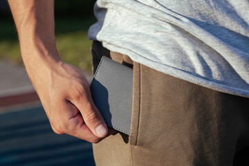 A young man in close-up sportswear puts a small and comfortable gray leather wallet in his back pocket against the background of the sky and trees. Training on the playground.  - obrazy, fototapety, plakaty