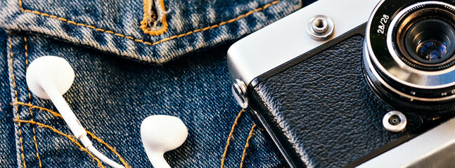 Film camera and white wired headphones lie against background of denim. Fashionable hipster...