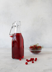 Red currant juice in a transparent bottle on a light table. Currant berries in a glass vase in the...