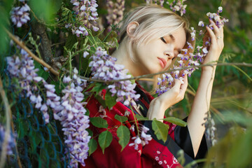 A beautiful girl with blonde hair in a traditional Chinese black and red dress. Portrait of a woman in a blooming wisteria garden, around flowers. The girl holds a branch of pink flowers in her hand