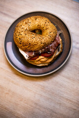 Bagel with cheese cream, dried tomato arugula, prosciutto ham, fresh tomato and balsamic cream on a plate on a wooden table.

