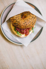 Bagel with cream cheese, beacon, pork, arugula, and tomato in a paper envelope on a plate on a wooden table.