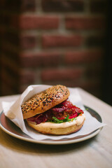 Bagel with cream cheese, beacon, pork, arugula, and tomato in a paper envelope on a plate on a wooden table against a brick wall.