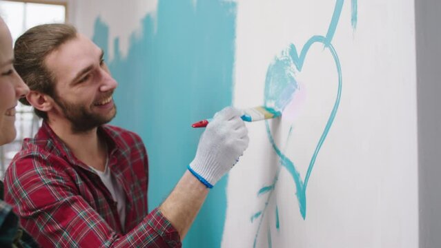 Good looking couple excited together painting walls at home in living room they make a big heart and painting with a small brushes together and smiling large
