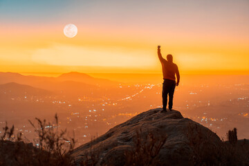 Plakat person standing on the top of the mountain with hand up, back view, over the city at sunset
