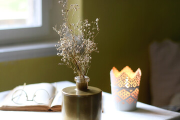 Open book, reading glasses, candle holder with lit candle and vase with gypsophila flowers. Dark background, selective focus.