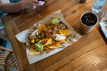 Plato de nachos con muchos condimentos en una mesa de madera y un refresco junto a la comida