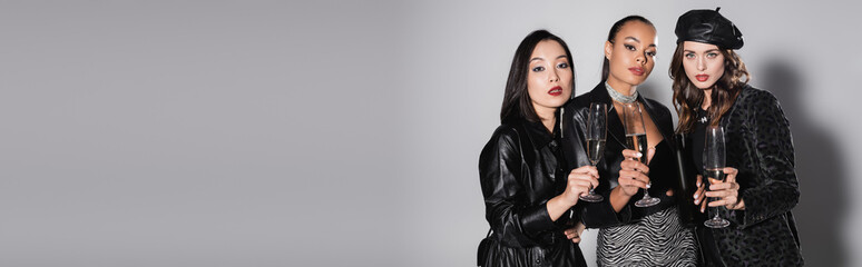 young multiethnic models holding glasses of champagne on grey, banner