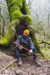 Chico con barba y equipado de senderismo descansando en el hueco de un arbol en un bosque con niebla