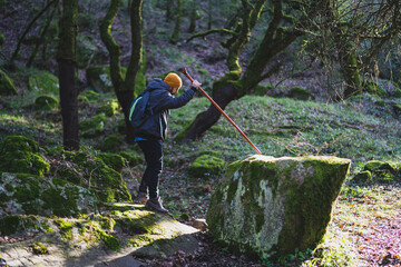 Chico joven con barba y ropa de abrigo montándose encima de una roca en ruta verde ayudado por un...