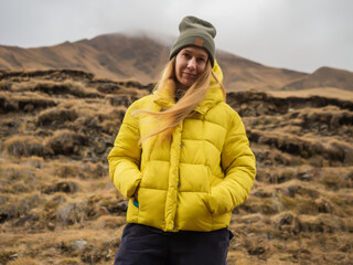 Naklejka premium A woman tourist in a quilted yellow jacket stands smiling against the backdrop of nature in the mountains