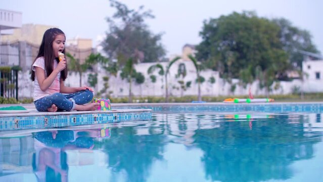 A young schoolgirl with Holi colors on her face and clothes enjoys eating ice cream in summers - Holi celebrations  good food. An adorable little kid eats an ice-cream cone while relaxing by the po...