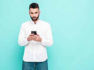 Handsome confident model.Sexy stylish man dressed in shirt and jeans. Fashion hipster male posing near blue wall in studio. Holding smartphone. Looking at cellphone screen. Using apps
