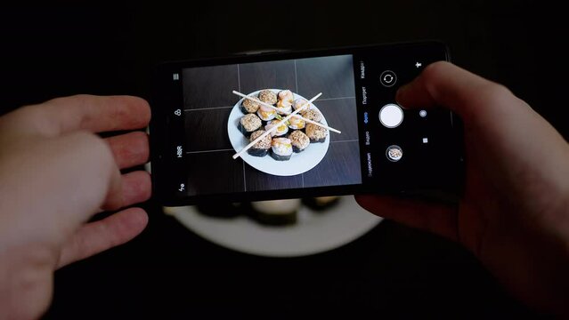 Female Hands Taking Photos of Prepared Food on a Smartphone on Table. Girl blogger shoots a picture, a photo, video review of sushi rolls on a mobile phone camera. Photographing food. Home kitchen.