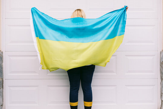 Ukrainian blonde-haired mature woman from the back with waving blue and yellow Ukrainian flag on white background in the street. Ukraine war, invasion, protest, patriotism and no war concept.