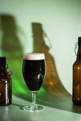 Glass of dark beer with foam head and empty bottles on colorful green and orange background, bright shadows