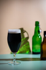 Glass of dark beer with foam head and empty bottles on colorful green and orange background, bright shadows