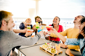Happy people toasting multi colored drinks at rooftop bar front of sea - Young friends having fun together drinking cocktails at happy hour at restaurant terrace - Social gathering party time concept