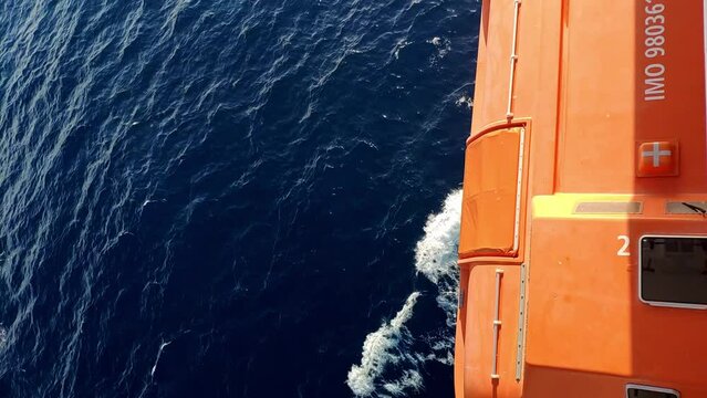 Vertical view of the side of a cruiser while navigating during the day. Orange life boat above sea water and waves on camera. Bright sunny day. Sailing is making typical waves to the sides.
