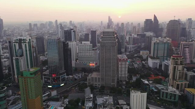 Drone descending over skyline, buildings and skyscrapers of Sukhumvit district during colorful warm sunrise in Bangkok, Thailand.