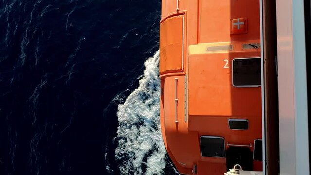Vertical view of the side of a cruiser while navigating. Orange life boat above sea water and waves on camera. Sailing is making typical waves to the sides. Nice evening sun light.