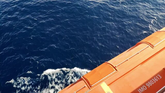 View of the sea while navigating on a cruiser. Orange life boat above  sea water on camera. Sailing is making typical small waves to the sides. Nice evening sun light.