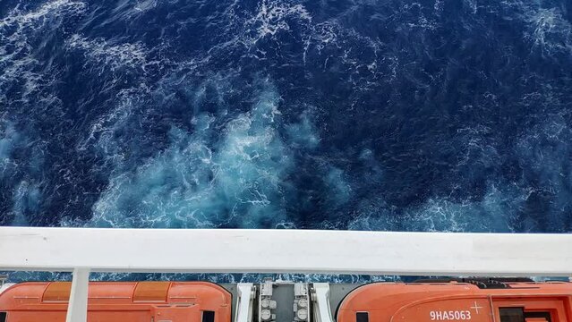 Sea water to the sides of a cruiser while navigating. Orange life boats hanging. Effects from engines propulsion can be seen on the water surface on the shape of white marks from air and small waves.