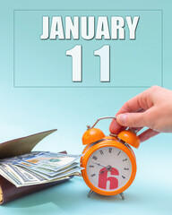 January 11th. Hand holding an orange alarm clock, a wallet with cash and a calendar date. Day 11 of month.