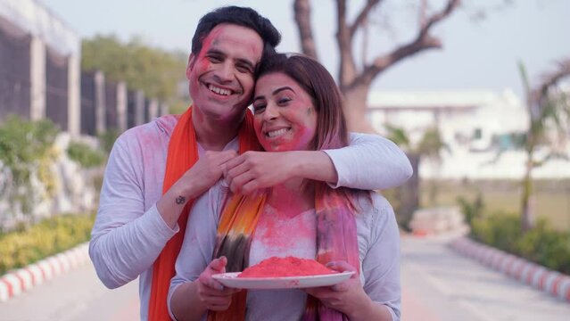 An attractive Indian couple standing together with a plate of bright colored Gulal - Holi celebrations  greetings  togetherness and bonding. Two youngsters standing in a public park with their face...