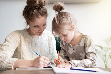 Naklejka na ściany i meble Caring young mother teaching little girl child cute daughter spelling, writing, sitting at desk, doing homework for school preparation. Family enjoying leisure time together at home.