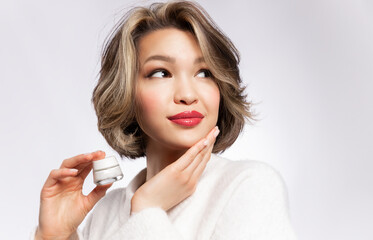 Pretty  girl holding mockup small cosmetic jar of rejuvenating serum or cream for skin around eyes posing on white studio background. Skin care cosmetics for natural beauty, advertisement.