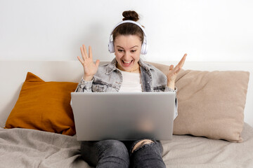 Young caucasian woman with headphones sitting on the bed with laptop computer. Girl using notebook...