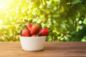 White bowl of fresh strawberries on a wooden table set outside against a background of green leaves