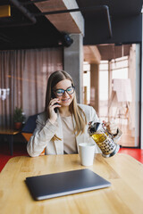 A modern woman of Slavic appearance, a manager in a light jacket and glasses, a girl with a smile on her face, sits at a table in a cafe and drinks tea. Remote work