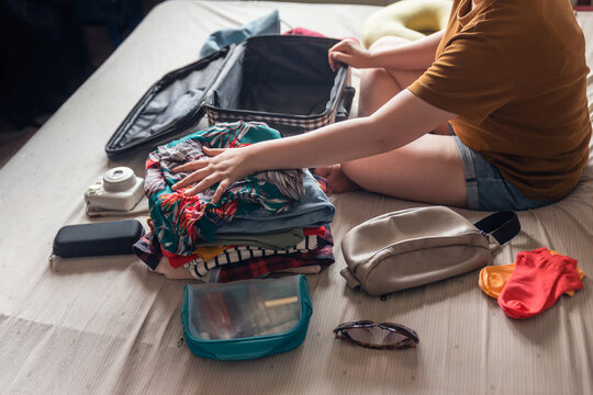 A woman folds clothes from a pile of selected items such as sunglasses, polaroid camera, cosmetic bag and small bag then puts them in luggage. Packing to go on a getaway vacation.