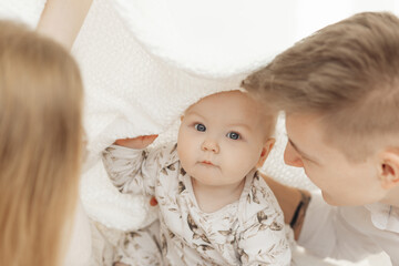 Portrait of beautiful smiling blue-eyed plump baby infant sitting between parents covered with white cotton blanket.