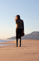 Backview of cowboy woman wears boots and brown hat. Flying hair in windy day. Sea background. Vertical photo.