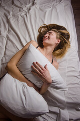 Beautiful thin young woman with long blonde hair in a white bed lies hugging a pillow and laughs