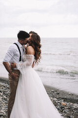 Fototapeta na wymiar The bride and groom in wedding dresses hug and walk together along the sea in nature on an outdoor trip. Husband and wife in love spend their honeymoon. Selective focus