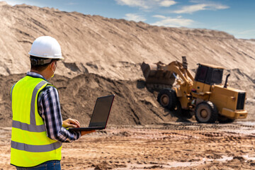 A Civil engineer using laptop to inspect work of Yellow Excavator working at sandpit. sand industry.
